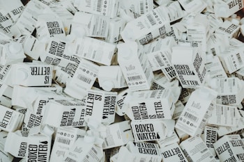 A large number of white cartons with black text saying 'Boxed Water is Better' are piled together. The cartons have graphics and text promoting sustainability.