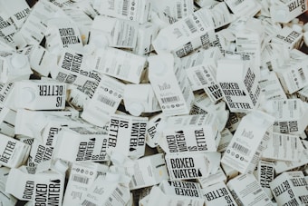 A large number of white cartons with black text saying 'Boxed Water is Better' are piled together. The cartons have graphics and text promoting sustainability.