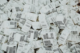 A large number of white cartons with black text saying 'Boxed Water is Better' are piled together. The cartons have graphics and text promoting sustainability.