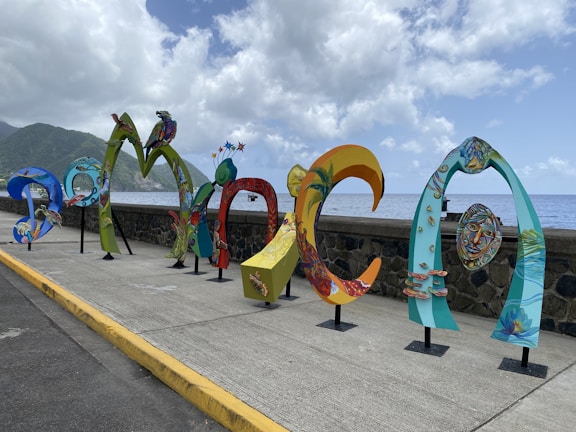 Colorful, artistic letters forming the word 'DOMINICA' are displayed along a seaside promenade. The letters are uniquely styled with vibrant patterns and illustrations, showcasing various elements of Dominican culture and nature. In the background, the ocean extends to the horizon under a partly cloudy sky, with lush green hills visible on the left side.