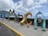 Colorful, artistic letters forming the word 'DOMINICA' are displayed along a seaside promenade. The letters are uniquely styled with vibrant patterns and illustrations, showcasing various elements of Dominican culture and nature. In the background, the ocean extends to the horizon under a partly cloudy sky, with lush green hills visible on the left side.