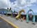 Colorful, artistic letters forming the word 'DOMINICA' are displayed along a seaside promenade. The letters are uniquely styled with vibrant patterns and illustrations, showcasing various elements of Dominican culture and nature. In the background, the ocean extends to the horizon under a partly cloudy sky, with lush green hills visible on the left side.