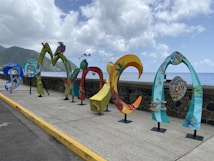 Colorful, artistic letters forming the word 'DOMINICA' are displayed along a seaside promenade. The letters are uniquely styled with vibrant patterns and illustrations, showcasing various elements of Dominican culture and nature. In the background, the ocean extends to the horizon under a partly cloudy sky, with lush green hills visible on the left side.
