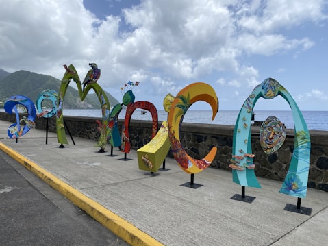 Colorful, artistic letters forming the word 'DOMINICA' are displayed along a seaside promenade. The letters are uniquely styled with vibrant patterns and illustrations, showcasing various elements of Dominican culture and nature. In the background, the ocean extends to the horizon under a partly cloudy sky, with lush green hills visible on the left side.