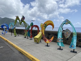 Colorful, artistic letters forming the word 'DOMINICA' are displayed along a seaside promenade. The letters are uniquely styled with vibrant patterns and illustrations, showcasing various elements of Dominican culture and nature. In the background, the ocean extends to the horizon under a partly cloudy sky, with lush green hills visible on the left side.