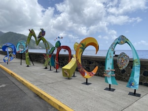 Colorful, artistic letters forming the word 'DOMINICA' are displayed along a seaside promenade. The letters are uniquely styled with vibrant patterns and illustrations, showcasing various elements of Dominican culture and nature. In the background, the ocean extends to the horizon under a partly cloudy sky, with lush green hills visible on the left side.