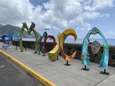 Colorful, artistic letters forming the word 'DOMINICA' are displayed along a seaside promenade. The letters are uniquely styled with vibrant patterns and illustrations, showcasing various elements of Dominican culture and nature. In the background, the ocean extends to the horizon under a partly cloudy sky, with lush green hills visible on the left side.