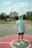 A young person stands on a basketball court, wearing a light blue cap, matching t-shirt, dark shorts, and sneakers. In the background, there are trees, a basketball hoop, and suburban houses under a partly cloudy sky.