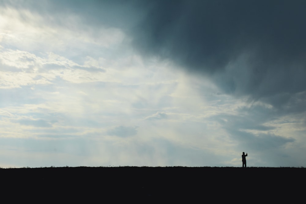 silhouette of person standing on top of hill under white clouds and blue sky during daytime