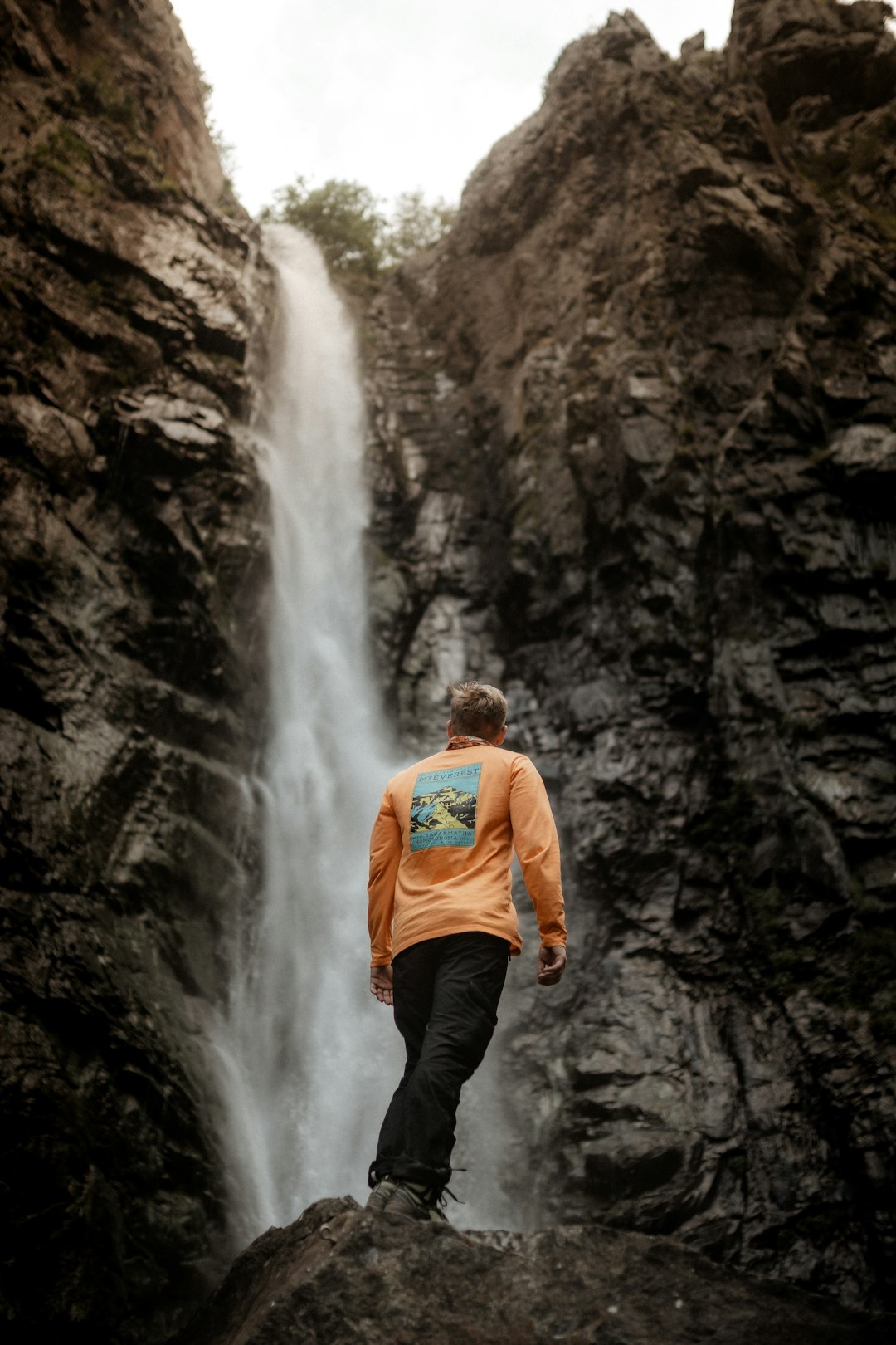 man in orange hoodie standing in front of waterfalls during daytime