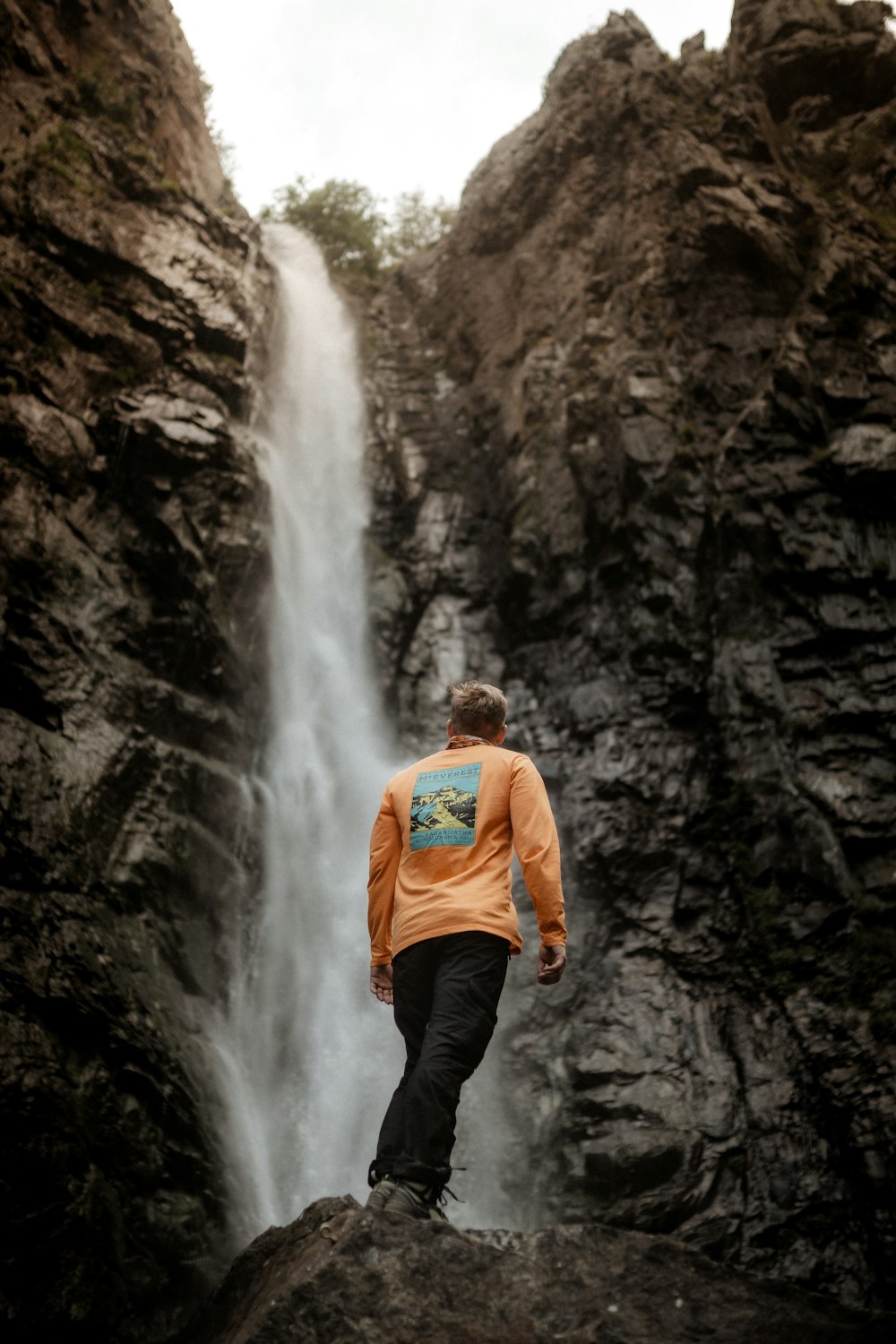 homme en sweat à capuche orange debout devant des chutes d’eau pendant la journée