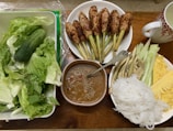 An assortment of fresh vegetables including lettuce and cucumber is placed in a container next to a bowl of grilled lemongrass chicken skewers. There is a bowl of light brown dipping sauce with a sprinkling of sesame seeds and chili pieces. Another bowl contains sliced cucumbers, eggplant, pineapple, and a portion of white rice noodles. A mug with a red pattern is also present.