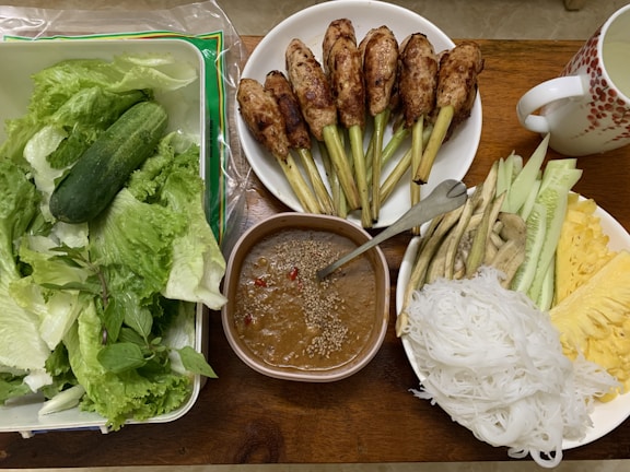 An assortment of fresh vegetables including lettuce and cucumber is placed in a container next to a bowl of grilled lemongrass chicken skewers. There is a bowl of light brown dipping sauce with a sprinkling of sesame seeds and chili pieces. Another bowl contains sliced cucumbers, eggplant, pineapple, and a portion of white rice noodles. A mug with a red pattern is also present.