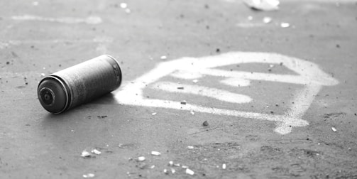 A spray paint can is lying on a surface next to a graffiti symbol that resembles a stylized eye or abstract pattern. The environment appears to be outdoors and rough, with scattered small debris around.