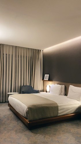A modern hotel room with soft ambient lighting, motorized blinds, and a bedside control panel