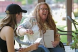 Two people are engaged in a lively conversation outdoors. One person, with long hair and glasses, is laughing and holding a notebook. The other person, wearing a black tank top and a black cap, is facing them and holding a notebook or paper.