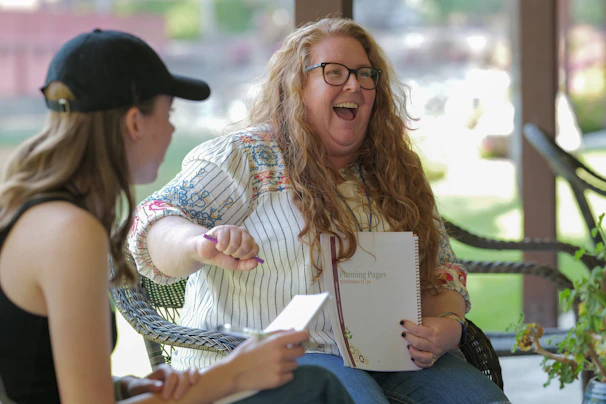 Two people are engaged in a lively conversation outdoors. One person, with long hair and glasses, is laughing and holding a notebook. The other person, wearing a black tank top and a black cap, is facing them and holding a notebook or paper.