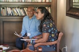 Two individuals are seated in a room filled with books, engaging in what appears to be a discussion or meeting. One person is wearing a light blue shirt and glasses, while the other has long hair and is dressed in a dark shirt with a tropical pattern. Both have papers in their hands and appear focused.