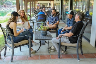 A group of people are sitting outside in a relaxed setting, engaging in conversation. The scene is on a patio with comfortable chairs and a table, surrounded by greenery and a reflective pond in the background. The group appears to be laughing and enjoying each other's company.