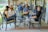 A group of people are sitting outside in a relaxed setting, engaging in conversation. The scene is on a patio with comfortable chairs and a table, surrounded by greenery and a reflective pond in the background. The group appears to be laughing and enjoying each other's company.