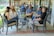 A group of people are sitting outside in a relaxed setting, engaging in conversation. The scene is on a patio with comfortable chairs and a table, surrounded by greenery and a reflective pond in the background. The group appears to be laughing and enjoying each other's company.