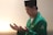 A man dressed in a green jacket and wearing a black cap is standing with his eyes closed and hands raised, as if in prayer or meditation. The background is softly lit, creating a calm atmosphere.