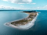 An aerial view of a narrow, elongated peninsula extending into the sea. The land is covered with patches of green grass and sandy beaches. The calm sea surrounds the peninsula, reflecting a clear blue sky with clouds scattered across.