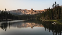 A tranquil lake with a clear reflection of the surrounding pine trees and distant mountains under a calm, early evening sky. The sunlight casts a warm glow on the rugged mountain peaks, while the still water mirrors the serene landscape.