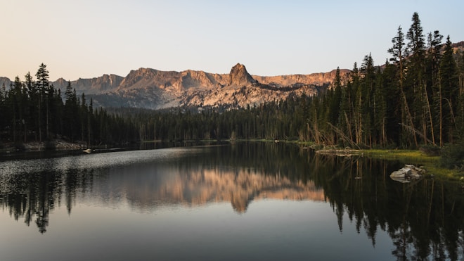A tranquil lake with a clear reflection of the surrounding pine trees and distant mountains under a calm, early evening sky. The sunlight casts a warm glow on the rugged mountain peaks, while the still water mirrors the serene landscape.
