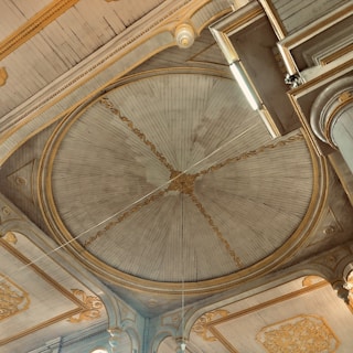 An ornate interior ceiling features a large, circular centerpiece with intricate carvings and golden accents. The ceiling is wooden, with a radial pattern emanating from the center. Surrounding the circle are decorative moldings and arches, all with detailed embellishments.