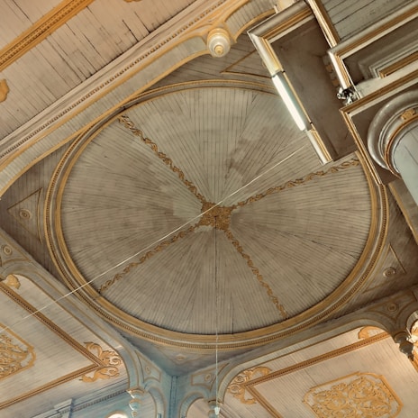 An ornate interior ceiling features a large, circular centerpiece with intricate carvings and golden accents. The ceiling is wooden, with a radial pattern emanating from the center. Surrounding the circle are decorative moldings and arches, all with detailed embellishments.