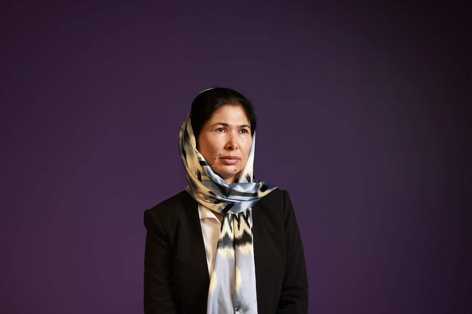 A woman in a dark blazer and patterned headscarf stands with a composed, confident expression against a deep purple background.