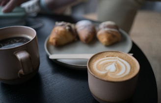 Two cups of coffee, one with latte art, are placed on a dark wooden table beside a plate holding three pastries. A wooden stir stick rests on the plate.