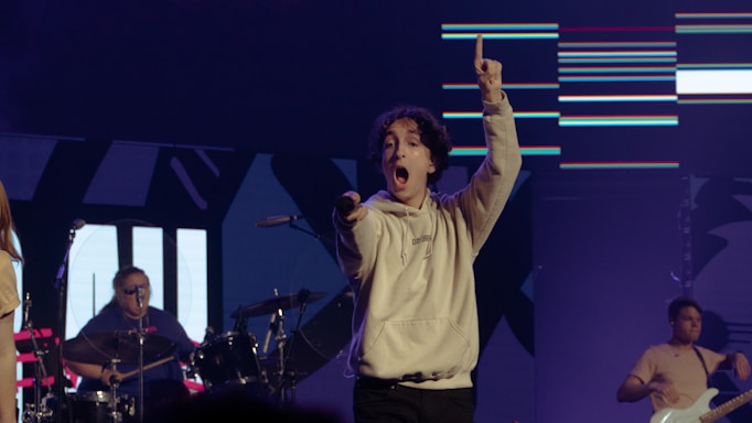 A musician energetically performs on stage, holding a microphone and pointing upwards. The drummer is visible in the background, playing the drums. Another musician with a guitar is also present, adding to the lively atmosphere. Bright lights and digital graphics are displayed on the backdrop, contributing to the concert setting.