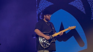 A person is playing an electric guitar, wearing a dark shirt and a cap, on a stage with a blue and black abstract design backdrop. The lighting is dim, creating a focused ambiance on the musician.