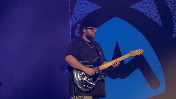 A person is playing an electric guitar, wearing a dark shirt and a cap, on a stage with a blue and black abstract design backdrop. The lighting is dim, creating a focused ambiance on the musician.