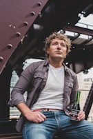 A person with curly hair is wearing a gray shirt over a white t-shirt and jeans, sitting on a metal structure. The individual is holding a green glass bottle of 'Sangría Señorial'. The background shows industrial elements and city buildings.