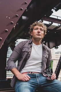 A person with curly hair is wearing a gray shirt over a white t-shirt and jeans, sitting on a metal structure. The individual is holding a green glass bottle of 'Sangría Señorial'. The background shows industrial elements and city buildings.