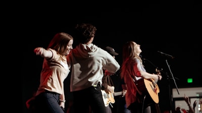 A dynamic and energetic scene with a group of young people on stage, engaged in a musical performance. One person is playing an acoustic guitar while others appear to be dancing and enjoying the atmosphere. The background is dark, adding focus to the well-lit performers.