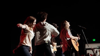 A dynamic and energetic scene with a group of young people on stage, engaged in a musical performance. One person is playing an acoustic guitar while others appear to be dancing and enjoying the atmosphere. The background is dark, adding focus to the well-lit performers.
