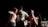 A dynamic and energetic scene with a group of young people on stage, engaged in a musical performance. One person is playing an acoustic guitar while others appear to be dancing and enjoying the atmosphere. The background is dark, adding focus to the well-lit performers.