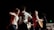 A dynamic and energetic scene with a group of young people on stage, engaged in a musical performance. One person is playing an acoustic guitar while others appear to be dancing and enjoying the atmosphere. The background is dark, adding focus to the well-lit performers.