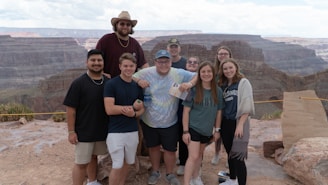 A group of eight young people are standing together outdoors at a scenic overlook with expansive canyon views in the background. They are casually dressed and smiling at the camera. The sky is partly cloudy, and the ground is rocky.