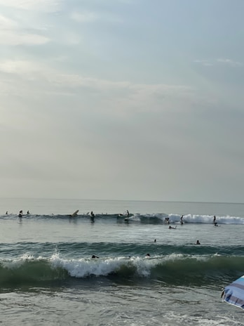 Several surfers are catching waves in the ocean near the shore under a cloudy sky. The waves appear moderate, with people riding on surfboards and others waiting for their turn in the water. A hint of a beach umbrella is visible in the corner, suggesting a leisurely beach setting.