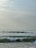 Several surfers are catching waves in the ocean near the shore under a cloudy sky. The waves appear moderate, with people riding on surfboards and others waiting for their turn in the water. A hint of a beach umbrella is visible in the corner, suggesting a leisurely beach setting.