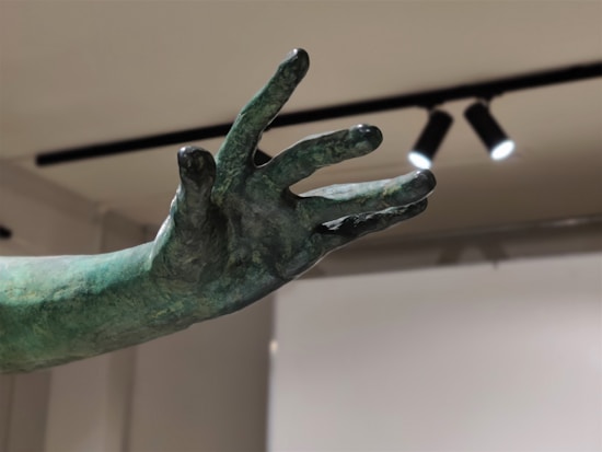 A detailed view of a greenish bronze sculpture hand, with fingers elegantly extended against a neutral background. The lighting above highlights the texture and contours of the hand.