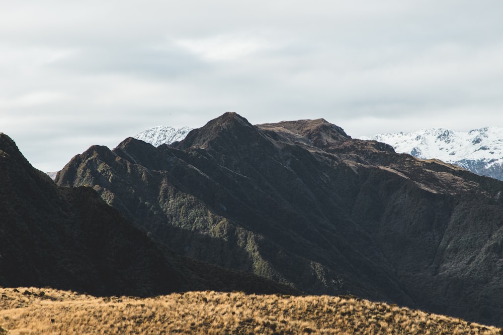 brown grass field near gray rocky mountain under white cloudy sky during daytime