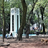 A park setting with tall green trees and a white clock tower with multiple clock faces. Several people are sitting on benches, some interacting and one accompanied by a dog. There are additional dogs roaming freely. The area is enclosed with a black fence, and the ground is covered with brown soil or mulch.
