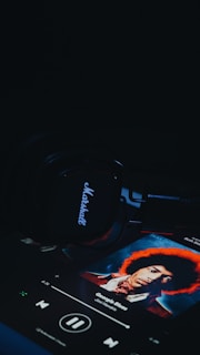 A set of Marshall headphones lies on a dark surface, with a music player screen displaying an album cover featuring a person with an afro hairstyle. The title on the screen reads 'George's Blues' by Jimi Hendrix.