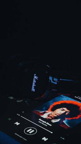 A set of Marshall headphones lies on a dark surface, with a music player screen displaying an album cover featuring a person with an afro hairstyle. The title on the screen reads 'George's Blues' by Jimi Hendrix.